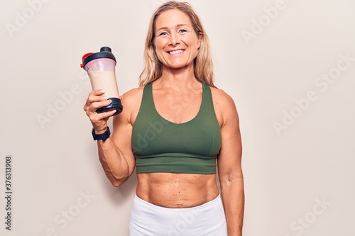 Middle age caucasian blonde woman wearing sport clothes drinking a protein shake looking positive and happy standing and smiling with a confident smile showing teeth