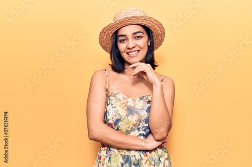 Young beautiful latin woman wearing summer hat looking confident at the camera smiling with crossed arms and hand raised on chin. thinking positive.