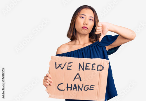 Young beautiful chinese girl holding we need a change banner with angry face, negative sign showing dislike with thumbs down, rejection concept