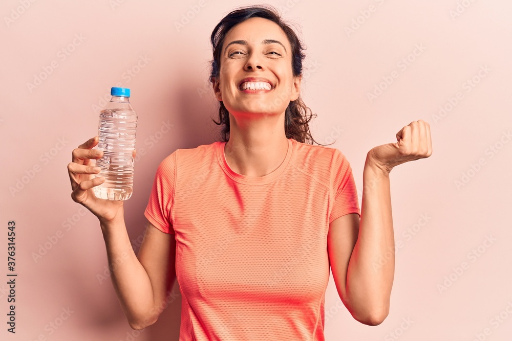 Young beautiful hispanic woman drinking bottle of water screaming proud, celebrating victory and success very excited with raised arm Young beautiful hispanic woman drinking bottle of water screaming proud, celebrating victory and success very excited with raised arm