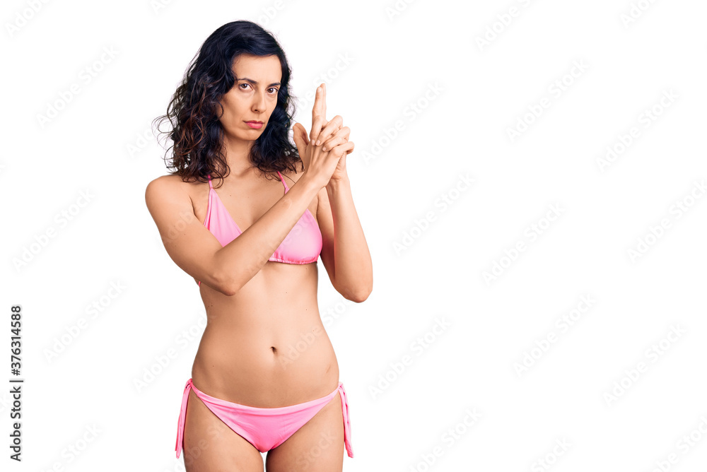 Young beautiful hispanic woman wearing bikini holding symbolic gun with hand gesture, playing killing shooting weapons, angry face Young beautiful hispanic woman wearing bikini holding symbolic gun with hand gesture, playing killing shooting weapons, angry face