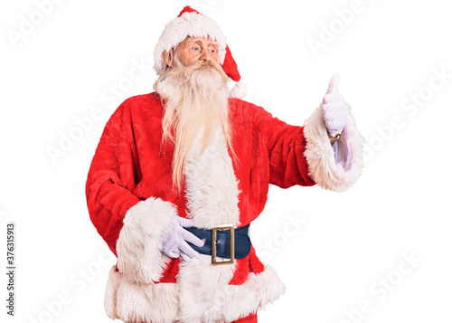 Old senior man with grey hair and long beard wearing traditional santa claus costume looking proud, smiling doing thumbs up gesture to the side
