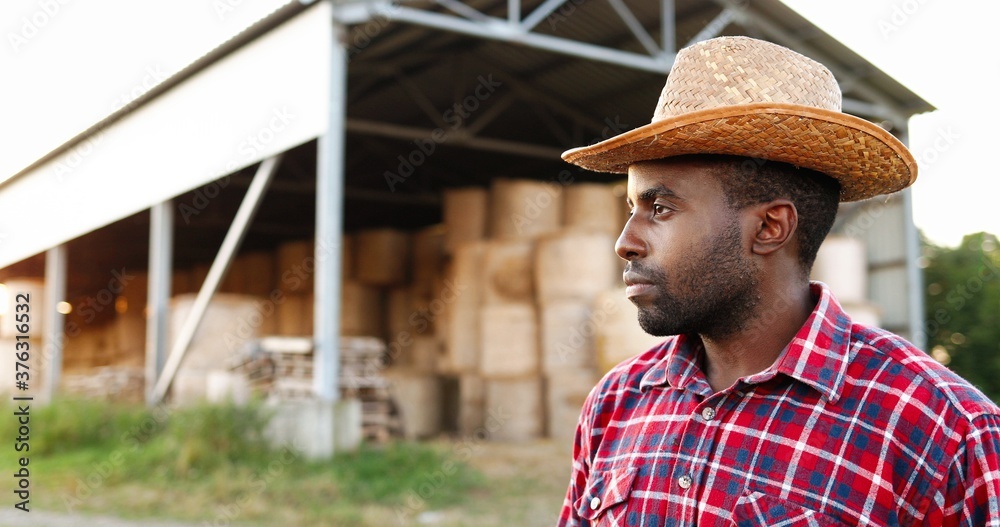 Young handsome African American man farmer in hat standing and smiling with shed full of hay on background. Portrait of happy cheerful male shepherd at stable. Outdoor. Close up.