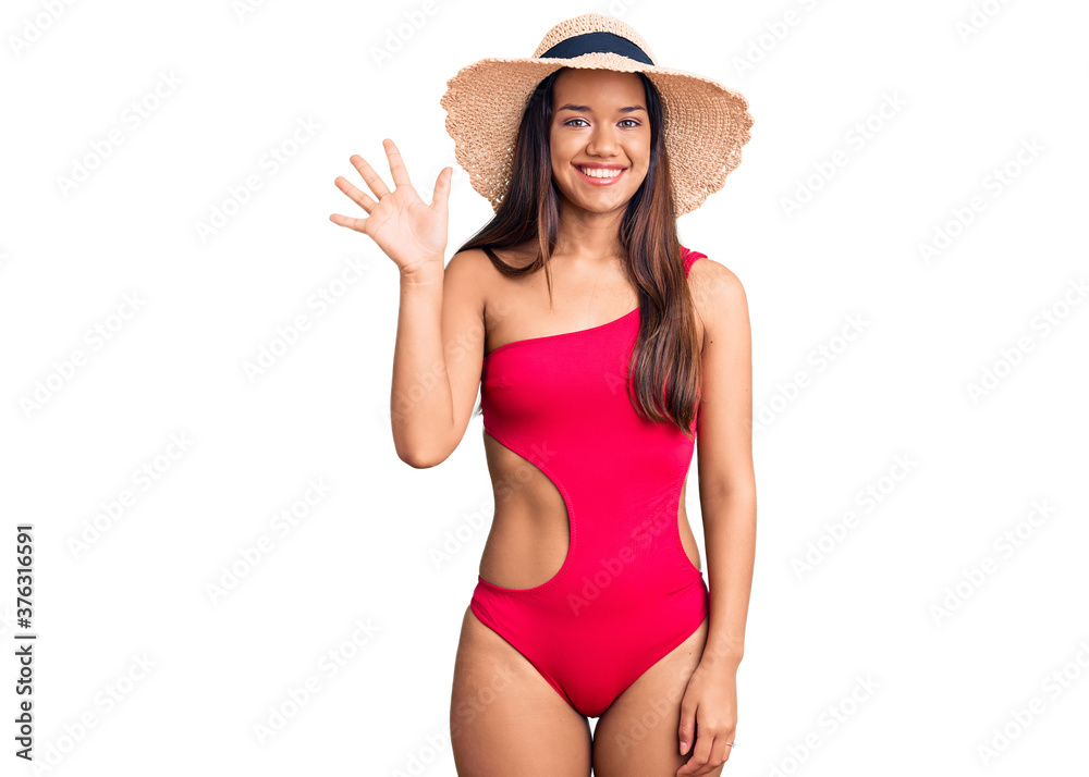 Young beautiful latin girl wearing swimwear and summer hat showing and pointing up with fingers number five while smiling confident and happy. Young beautiful latin girl wearing swimwear and summer hat showing and pointing up with fingers number five while smiling confident and happy.