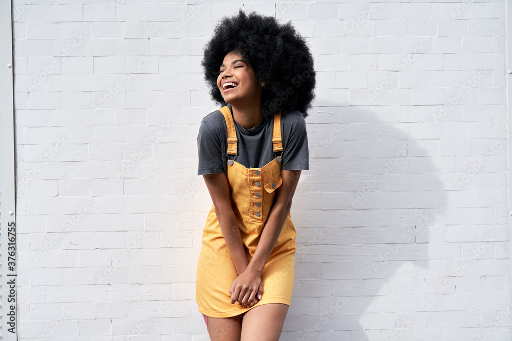 Happy stylish African American young woman wears yellow trendy sundress with Afro hair laughing looking away standing against white brick wall outdoor background. Smiling black hipster woman portrait - obrazy, fototapety, plakaty 