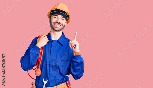 Young hispanic man wearing elecrician uniform holding cable surprised with an idea or question pointing finger with happy face, number one