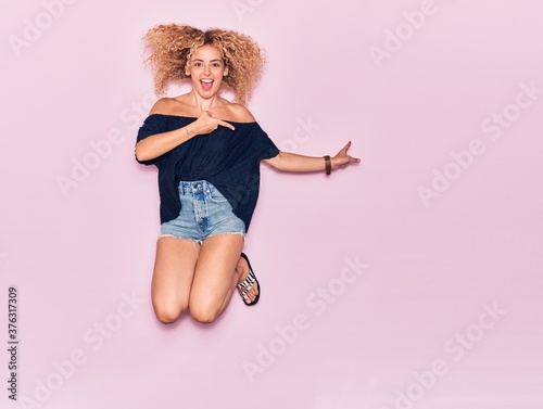 Young beautiful curly girl wearing casual clothes smiling happy. Jumping with smile on face pointing with fingers to the side over isolated pink background.
