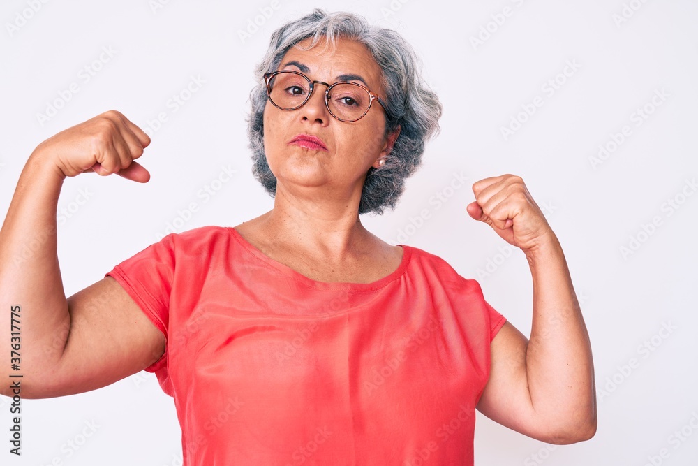 Senior hispanic grey- haired woman wearing casual clothes and glasses showing arms muscles smiling proud. fitness concept. Senior hispanic grey- haired woman wearing casual clothes and glasses showing arms muscles smiling proud. fitness concept.