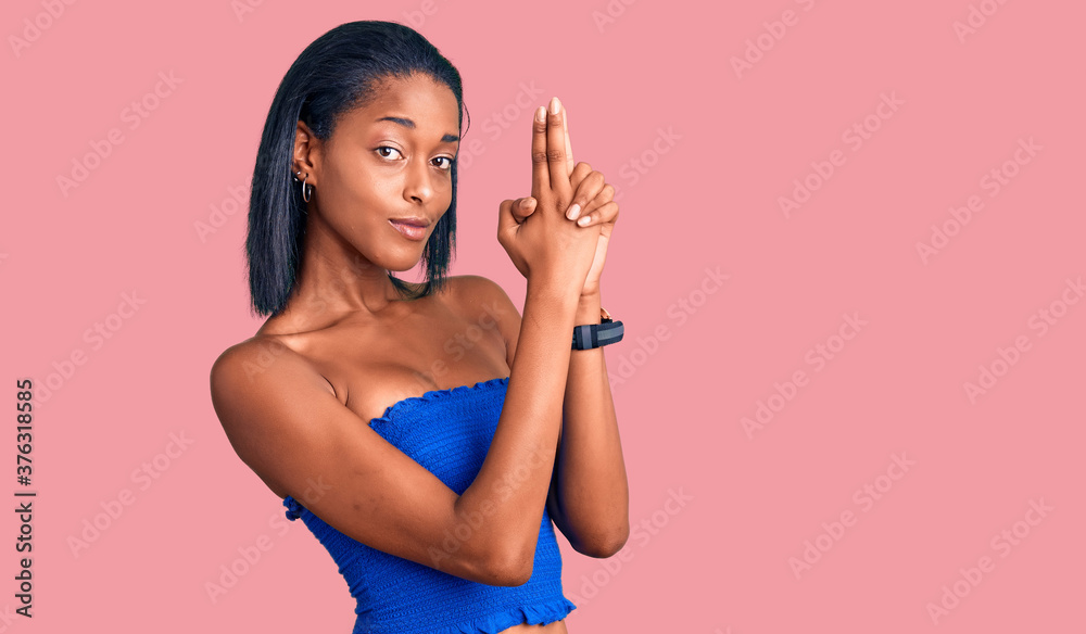 Fototapeta premium Young african american woman wearing casual summer clothes holding symbolic gun with hand gesture, playing killing shooting weapons, angry face
