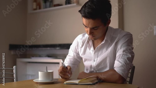 Dark-haired boy making notes in a notebook and drinking coffee. Teenager drawing and drinking something at wooden table. Man thinking and writing while using his white mug.
