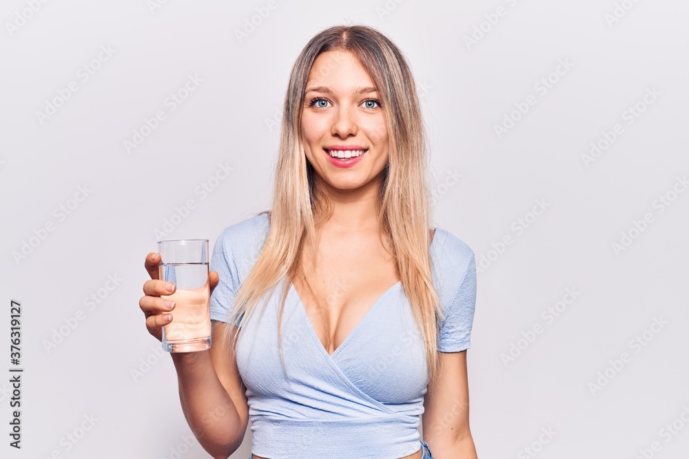 Young beautiful blonde girl drinking glass of water looking positive and happy standing and smiling with a confident smile showing teeth Young beautiful blonde girl drinking glass of water looking positive and happy standing and smiling with a confident smile showing teeth