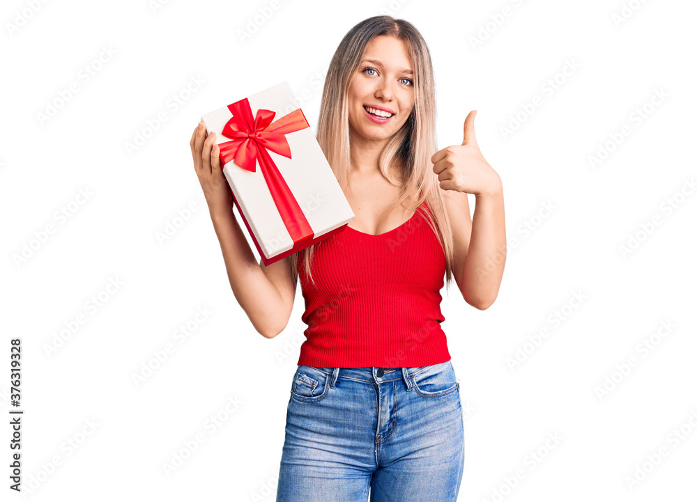Young beautiful blonde woman holding gift smiling happy and positive, thumb up doing excellent and approval sign Young beautiful blonde woman holding gift smiling happy and positive, thumb up doing excellent and approval sign