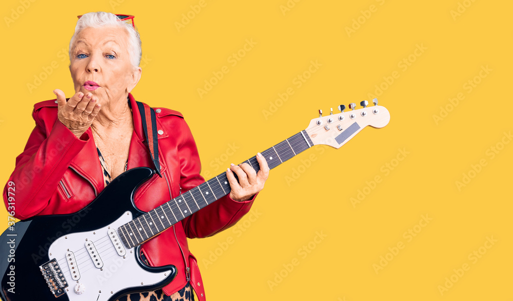Fototapeta premium Senior beautiful woman with blue eyes and grey hair wearing a modern look playing electric guitar looking at the camera blowing a kiss with hand on air being lovely and sexy. love expression.