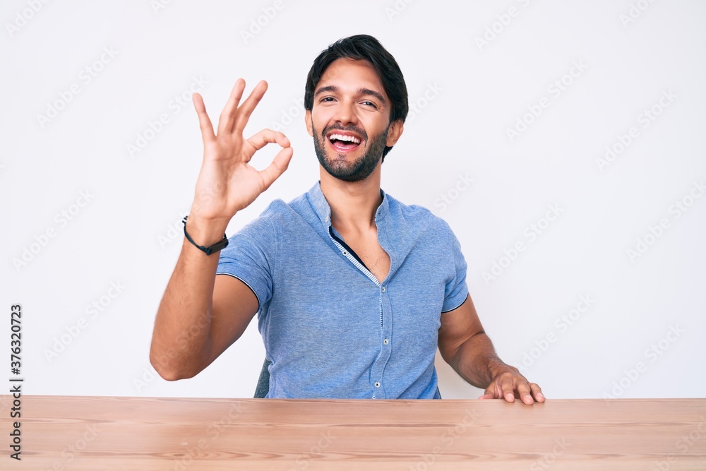 Handsome hispanic man wearing casual clothes sitting on the table smiling positive doing ok sign with hand and fingers. successful expression. Handsome hispanic man wearing casual clothes sitting on the table smiling positive doing ok sign with hand and fingers. successful expression.