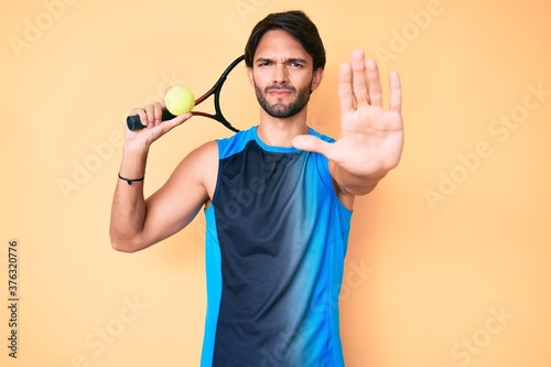 Handsome hispanic man playing tennis holding racket and ball with open hand doing stop sign with serious and confident expression, defense gesture