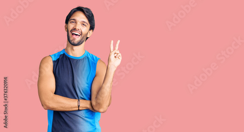 Handsome hispanic man wearing sportswear smiling with happy face winking at the camera doing victory sign. number two.
