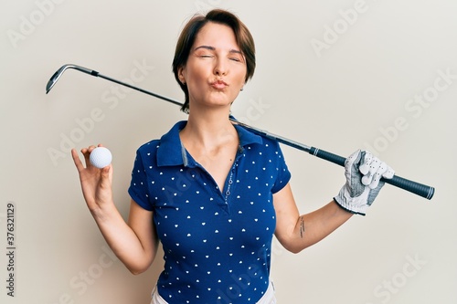 Young brunette woman with short hair holding ball and golf club looking at the camera blowing a kiss being lovely and sexy. love expression.