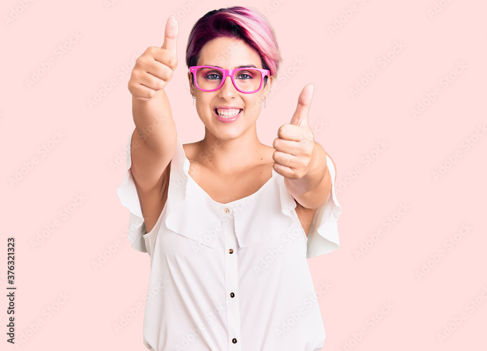 Young beautiful woman with pink hair wearing casual clothes and glasses approving doing positive gesture with hand, thumbs up smiling and happy for success. winner gesture. Young beautiful woman with pink hair wearing casual clothes and glasses approving doing positive gesture with hand, thumbs up smiling and happy for success. winner gesture.