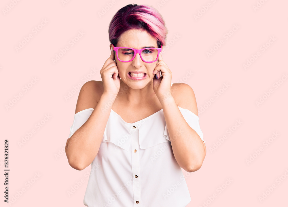 Young beautiful woman with pink hair wearing casual clothes and glasses covering ears with fingers with annoyed expression for the noise of loud music. deaf concept. Young beautiful woman with pink hair wearing casual clothes and glasses covering ears with fingers with annoyed expression for the noise of loud music. deaf concept.