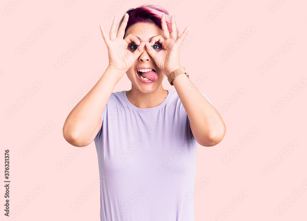 Young beautiful woman with pink hair wearing casual clothes doing ok gesture like binoculars sticking tongue out, eyes looking through fingers. crazy expression. Young beautiful woman with pink hair wearing casual clothes doing ok gesture like binoculars sticking tongue out, eyes looking through fingers. crazy expression.