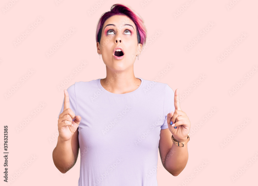 Young beautiful woman with pink hair wearing casual clothes amazed and surprised looking up and pointing with fingers and raised arms. Young beautiful woman with pink hair wearing casual clothes amazed and surprised looking up and pointing with fingers and raised arms.