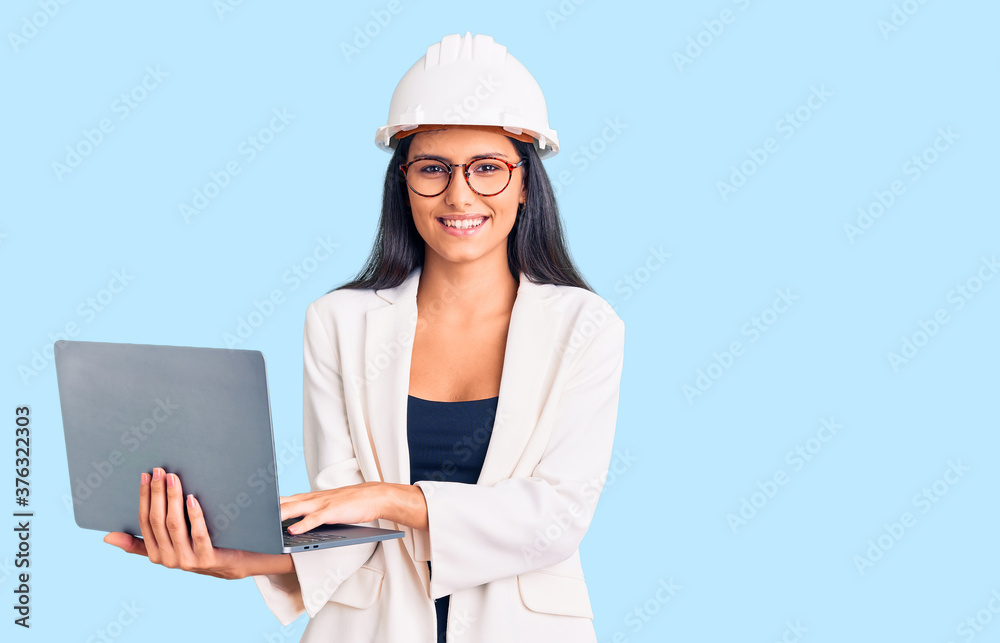 Young beautiful latin girl wearing architect hardhat holding laptop looking positive and happy standing and smiling with a confident smile showing teeth Young beautiful latin girl wearing architect hardhat holding laptop looking positive and happy standing and smiling with a confident smile showing teeth