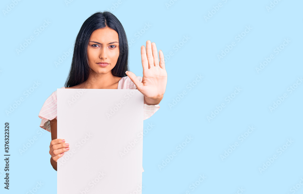 Young beautiful latin girl holding blank empty banner with open hand doing stop sign with serious and confident expression, defense gesture Young beautiful latin girl holding blank empty banner with open hand doing stop sign with serious and confident expression, defense gesture