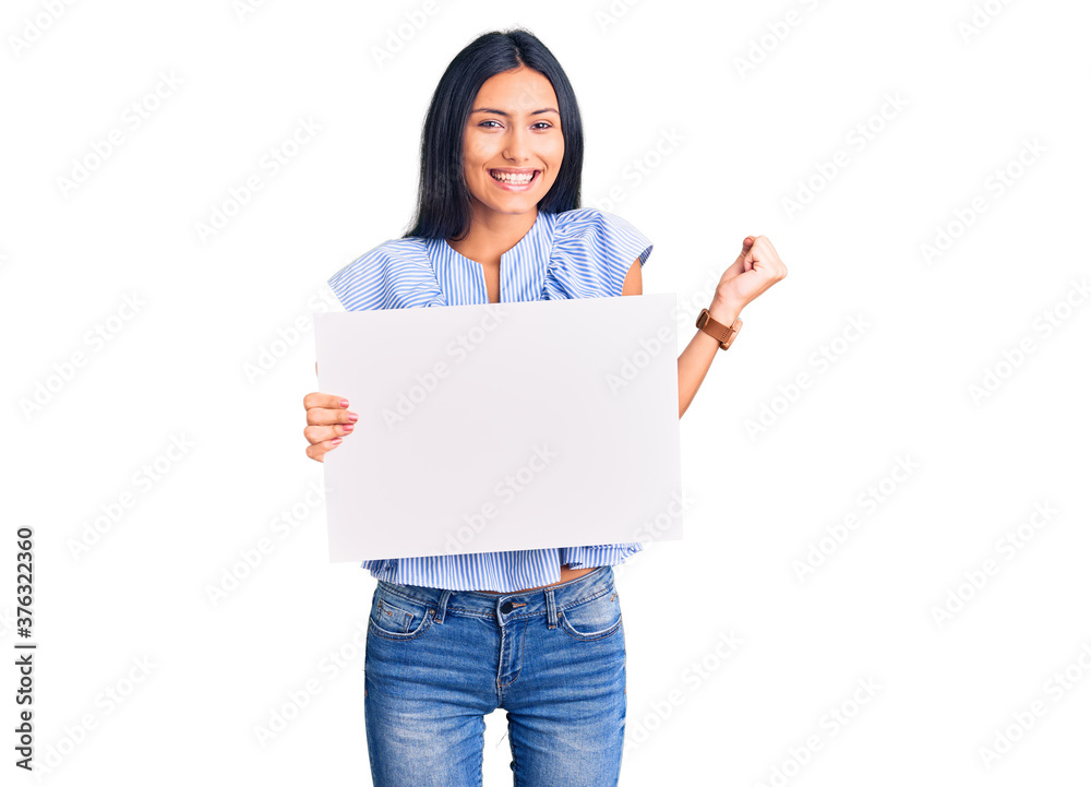 Young beautiful latin girl holding blank empty banner screaming proud, celebrating victory and success very excited with raised arms Young beautiful latin girl holding blank empty banner screaming proud, celebrating victory and success very excited with raised arms