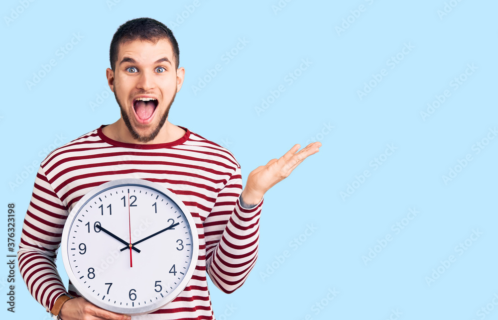 Young handsome man holding big clock celebrating victory with happy smile and winner expression with raised hands Young handsome man holding big clock celebrating victory with happy smile and winner expression with raised hands