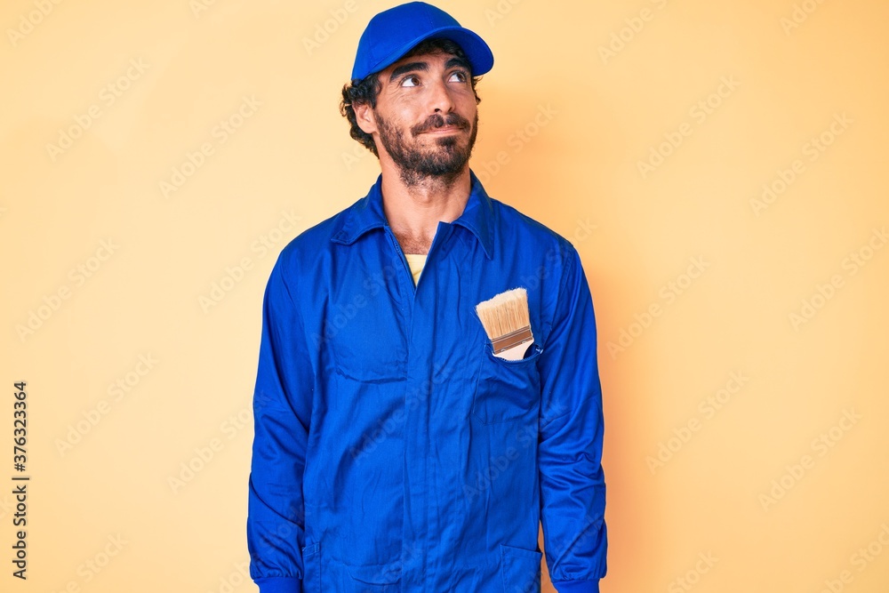 Handsome young man with curly hair and bear wearing builder jumpsuit uniform smiling looking to the side and staring away thinking.