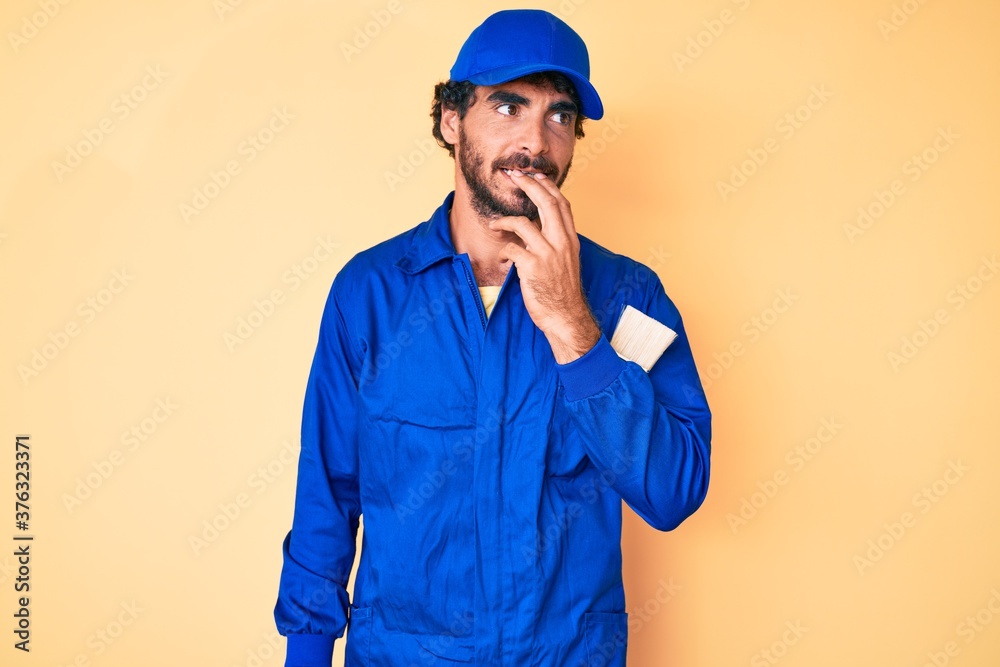 Handsome young man with curly hair and bear wearing builder jumpsuit uniform looking stressed and nervous with hands on mouth biting nails. anxiety problem.