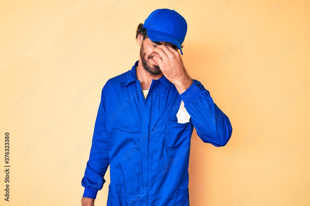 Handsome young man with curly hair and bear wearing builder jumpsuit uniform tired rubbing nose and eyes feeling fatigue and headache. stress and frustration concept.