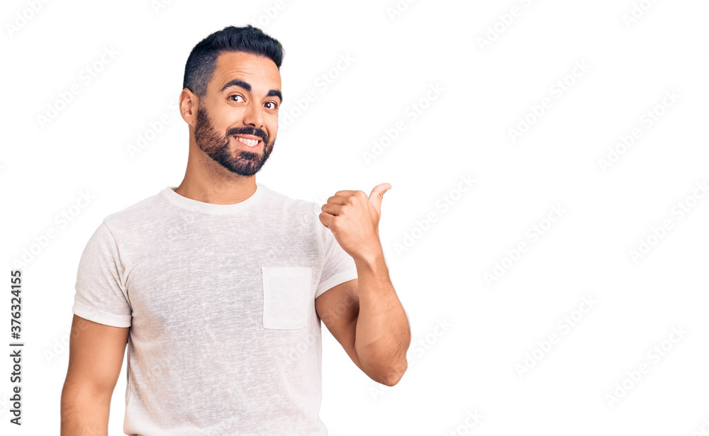 Young hispanic man wearing casual clothes smiling with happy face looking and pointing to the side with thumb up. Young hispanic man wearing casual clothes smiling with happy face looking and pointing to the side with thumb up.