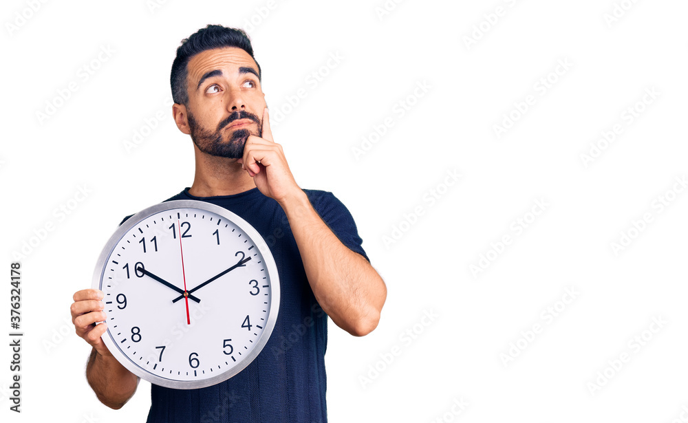 Young hispanic man holding big clock serious face thinking about question with hand on chin, thoughtful about confusing idea Young hispanic man holding big clock serious face thinking about question with hand on chin, thoughtful about confusing idea