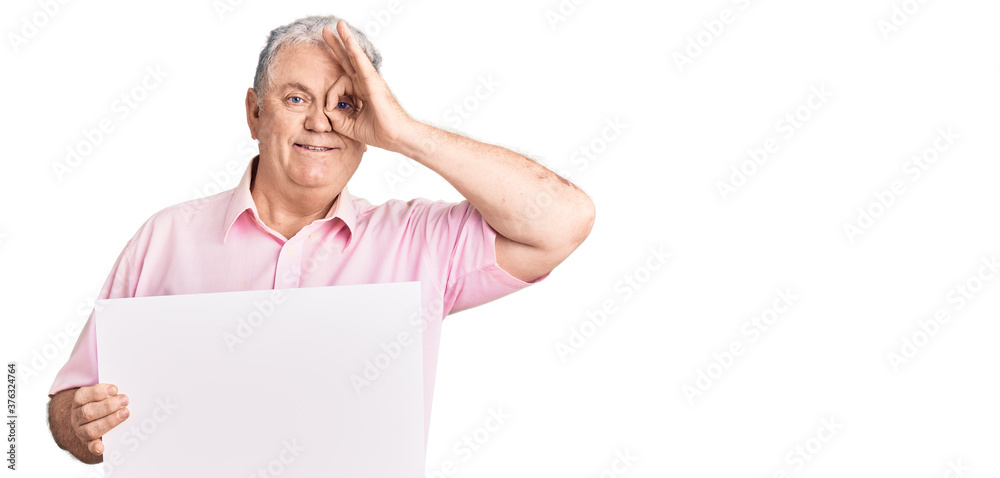 Senior grey-haired man holding blank empty banner smiling happy doing ok sign with hand on eye looking through fingers Senior grey-haired man holding blank empty banner smiling happy doing ok sign with hand on eye looking through fingers
