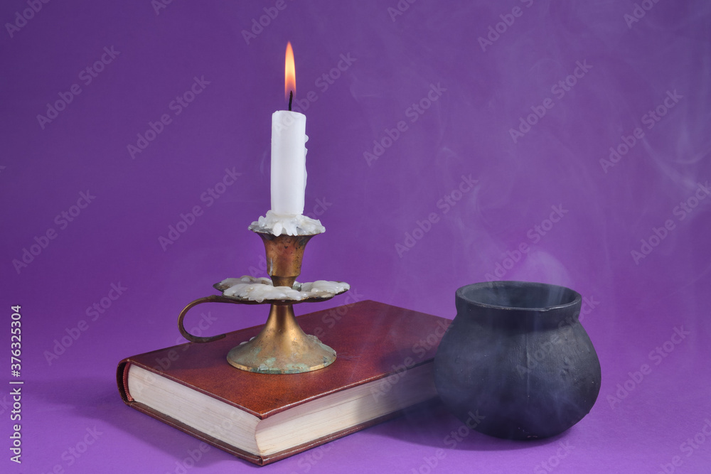 Candle in candlestick burning on an old book and witch smoky pot on the dark purple smoked background. Halloween concept Candle in candlestick burning on an old book and witch smoky pot on the dark purple smoked background. Halloween concept