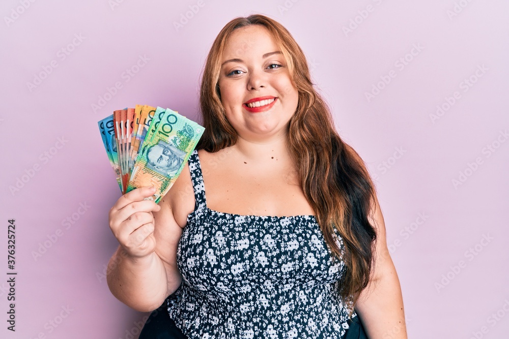 Plus size caucasian young woman holding australian dollars looking positive and happy standing and smiling with a confident smile showing teeth Plus size caucasian young woman holding australian dollars looking positive and happy standing and smiling with a confident smile showing teeth