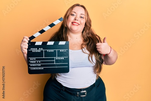 Plus size caucasian young woman holding video film clapboard smiling happy and positive  thumb up doing excellent and approval sign
