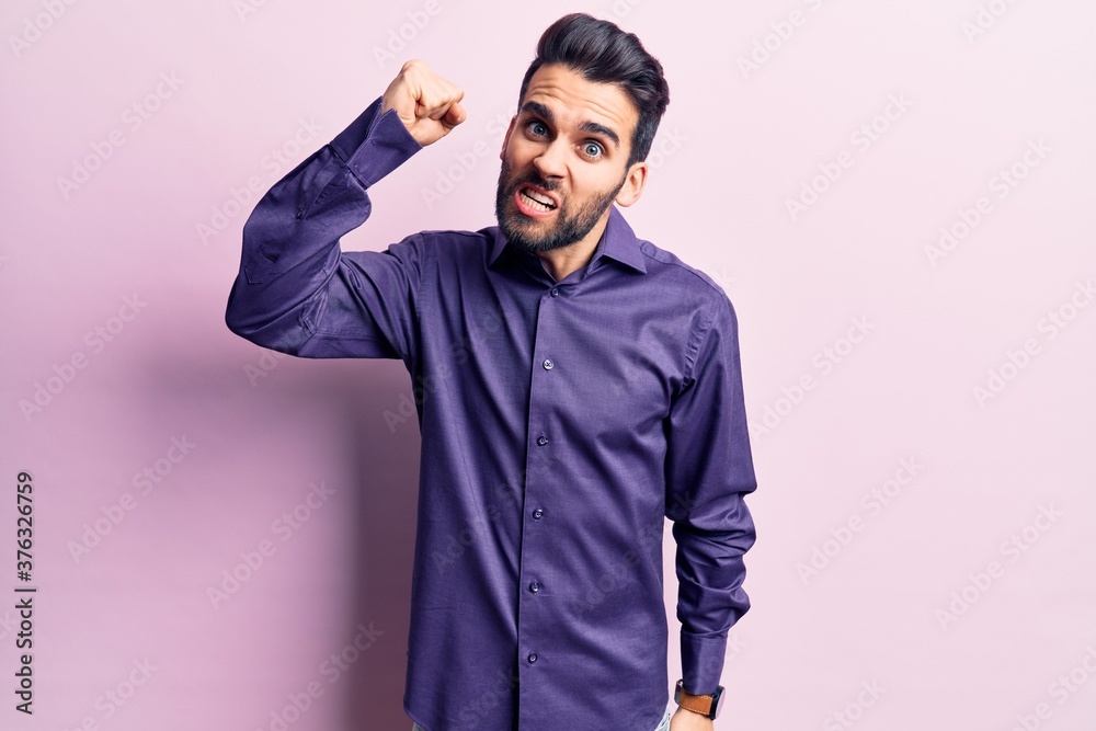 Young handsome man with beard wearing casual shirt angry and mad raising fist frustrated and furious while shouting with anger. rage and aggressive concept.