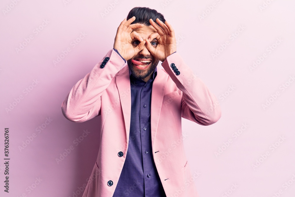 Young handsome man with beard wearing elegant jacket doing ok gesture like binoculars sticking tongue out, eyes looking through fingers. crazy expression. Young handsome man with beard wearing elegant jacket doing ok gesture like binoculars sticking tongue out, eyes looking through fingers. crazy expression.