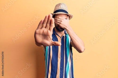 Young handsome man with beard wearing summer hat and shirt covering eyes with hands and doing stop gesture with sad and fear expression. embarrassed and negative concept.