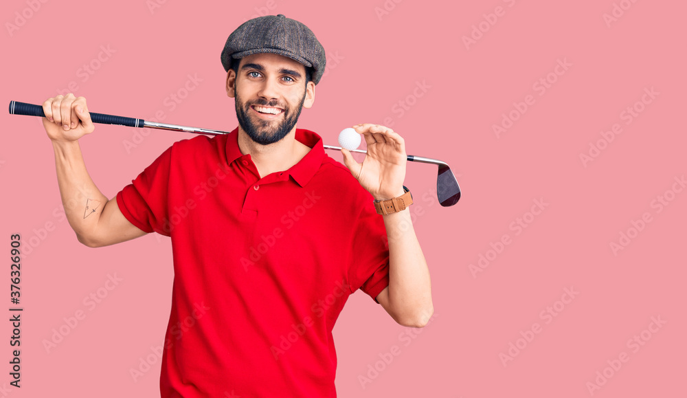 Young handsome man with beard playing golf holding club and ball looking positive and happy standing and smiling with a confident smile showing teeth Young handsome man with beard playing golf holding club and ball looking positive and happy standing and smiling with a confident smile showing teeth