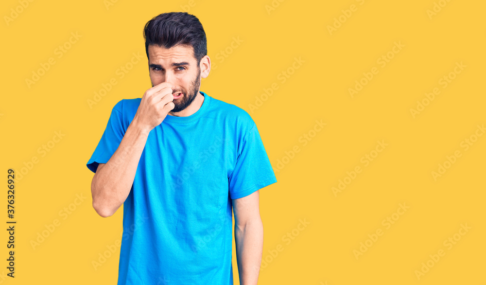 Young handsome man with beard wearing casual t-shirt smelling something stinky and disgusting, intolerable smell, holding breath with fingers on nose. bad smell Young handsome man with beard wearing casual t-shirt smelling something stinky and disgusting, intolerable smell, holding breath with fingers on nose. bad smell