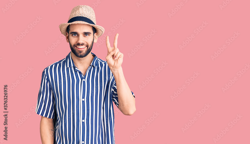 Young handsome man with beard wearing summer hat and striped shirt showing and pointing up with fingers number two while smiling confident and happy. Young handsome man with beard wearing summer hat and striped shirt showing and pointing up with fingers number two while smiling confident and happy.