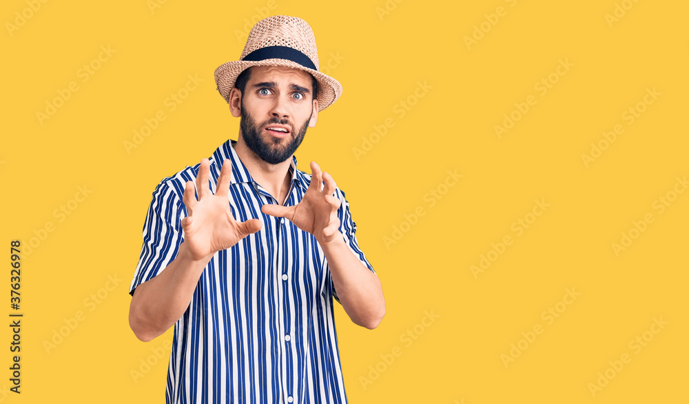 Young handsome man with beard wearing summer hat and striped shirt disgusted expression, displeased and fearful doing disgust face because aversion reaction. with hands raised