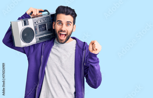 Young handsome man with beard listening to music using vintage boombox screaming proud, celebrating victory and success very excited with raised arms