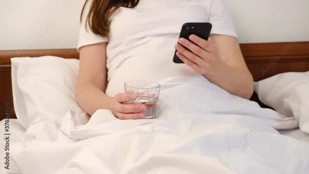Close up of unrecognizable young pregnant woman sit on white bed holding smartphone in hands and fresh water. Lady chatting in social networks, watching videos, using mobile applications at home Close up of unrecognizable young pregnant woman sit on white bed holding smartphone in hands and fresh water. Lady chatting in social networks, watching videos, using mobile applications at home