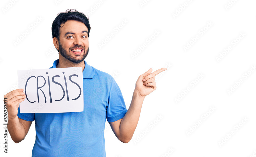 Young hispanic man holding crisis message paper smiling happy pointing with hand and finger to the side Young hispanic man holding crisis message paper smiling happy pointing with hand and finger to the side