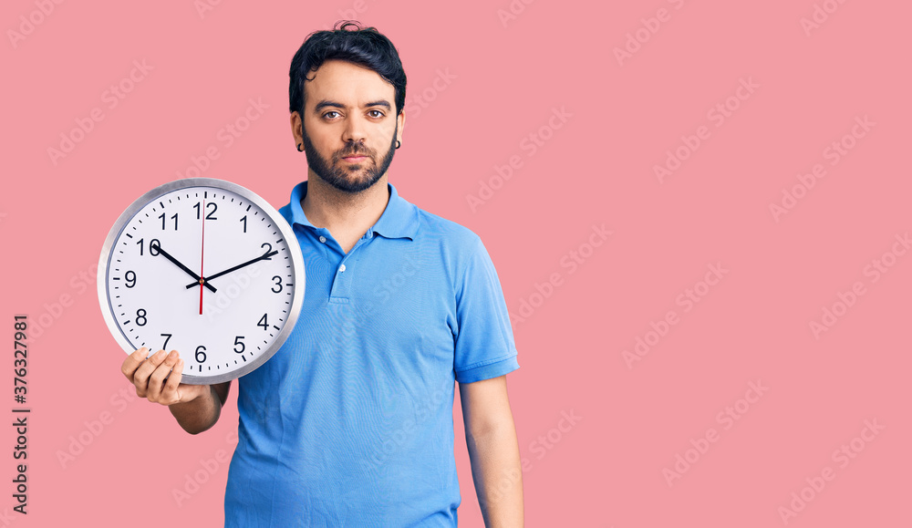 Young hispanic man holding big clock thinking attitude and sober expression looking self confident Young hispanic man holding big clock thinking attitude and sober expression looking self confident