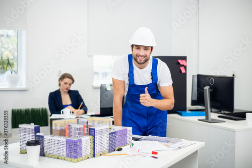 Young male architect working in the office. He is leaning on the table with a large construction maquette on it and showing thumbs up.